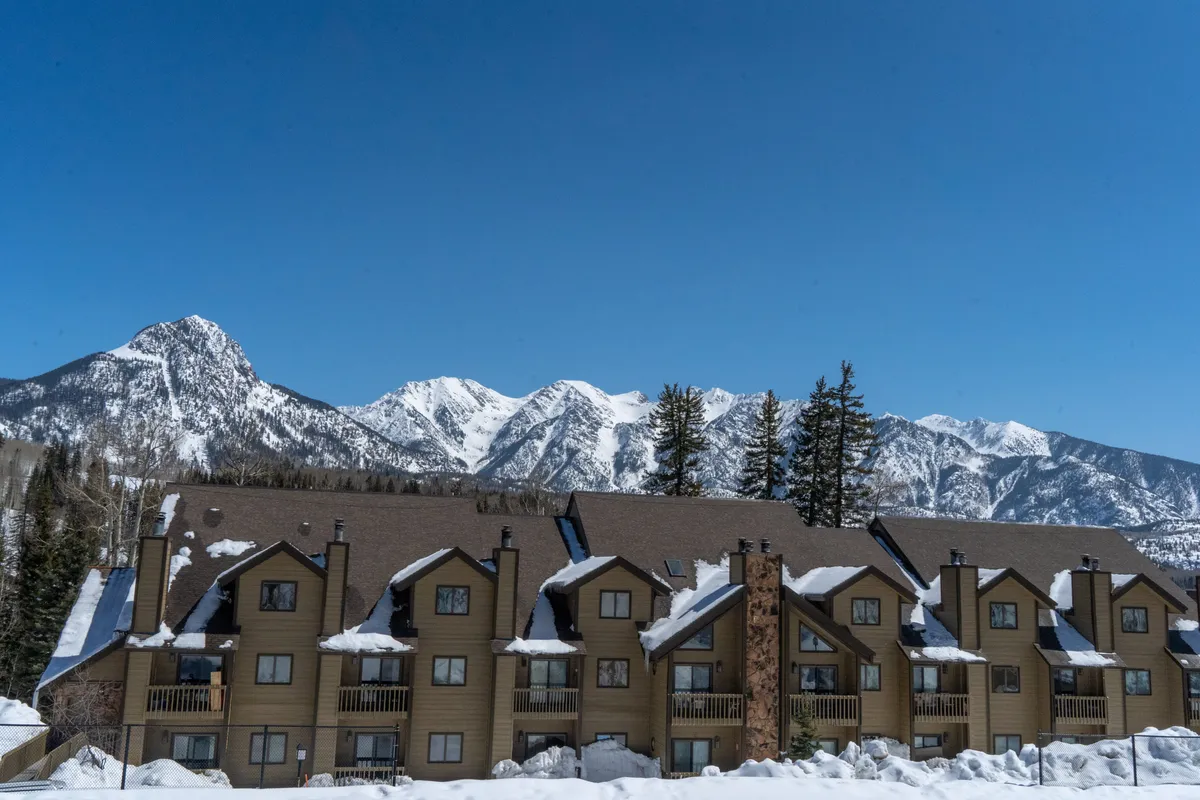 An exterior shot of a multi-level lodge set against snow-covered mountains and pine trees under a blue sky. The scene captures the adventurous spirit of the property and its proximity to outdoor activities.
