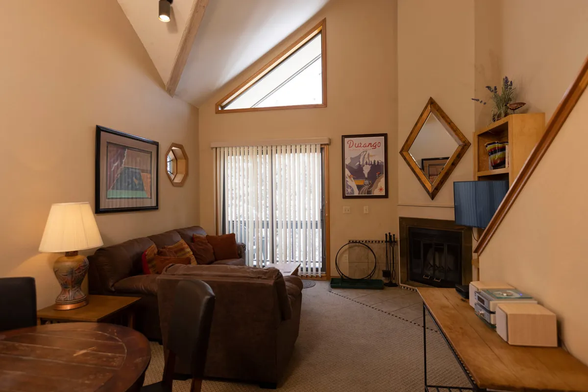 A cozy living room space featuring a comfortable sofa, a warm fireplace, artistic accents, and a large window framing the outdoors. The inviting seating arrangement is positioned for both relaxation and socializing, with natural light highlighting the mountain ambiance.