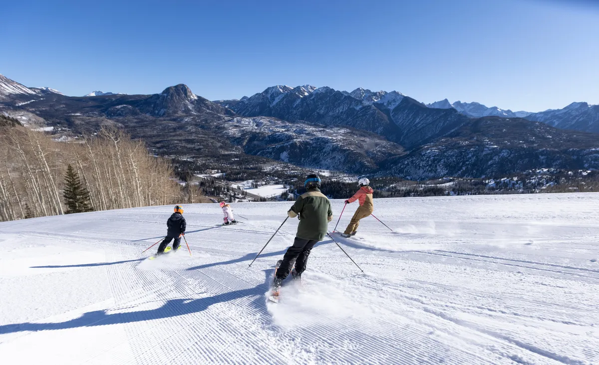A striking view of several skiers descending pristine, snow-covered slopes, with majestic mountains and a clear blue sky in the background. The image captures the thrill of skiing and the expansive beauty of the winter landscape.