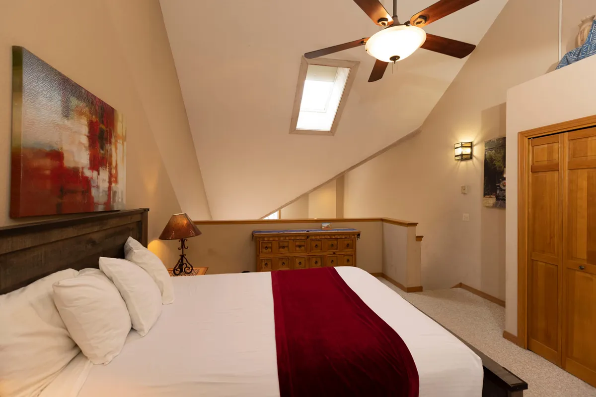 A sunlit bedroom featuring a cozy bed with a striking red accent, tasteful artwork on the wall, a wooden dresser, and a ceiling fan. The inviting atmosphere encourages relaxation, reading, or simply enjoying a peaceful start to the day.