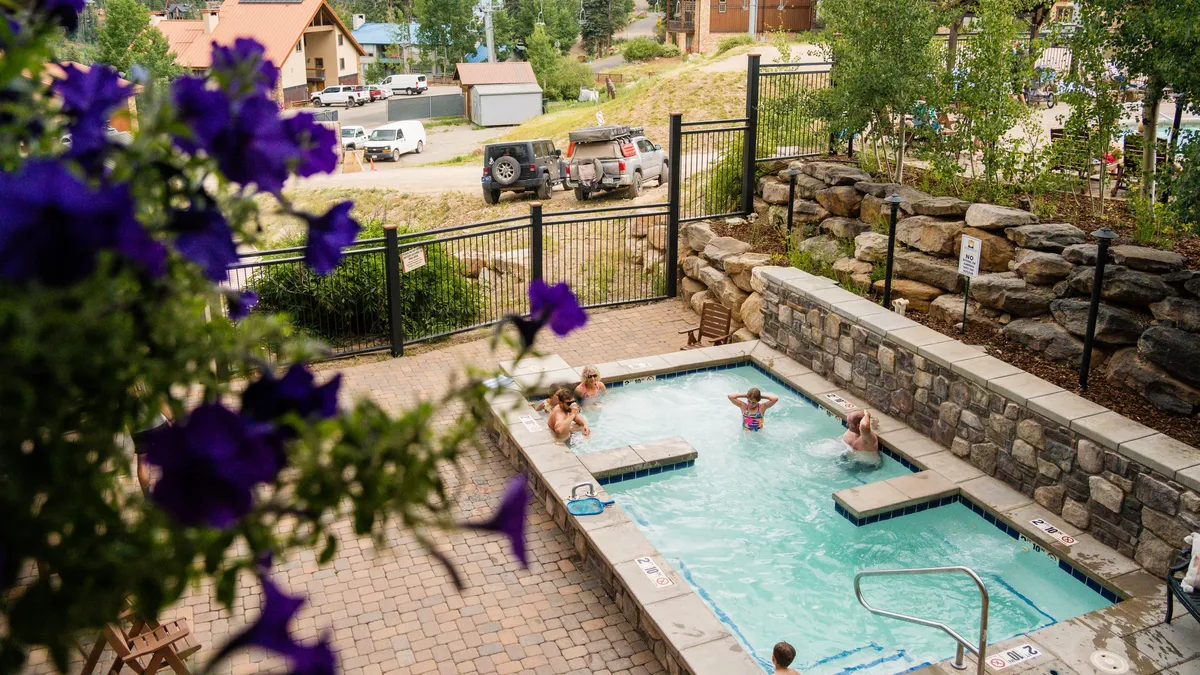 An exterior view highlighting a sparkling swimming pool and a bubbling hot tub set amidst flowering plants and a stone wall. The surrounding landscape gives a relaxed, outdoor resort feel, perfect for socializing or unwinding after a day of activity.