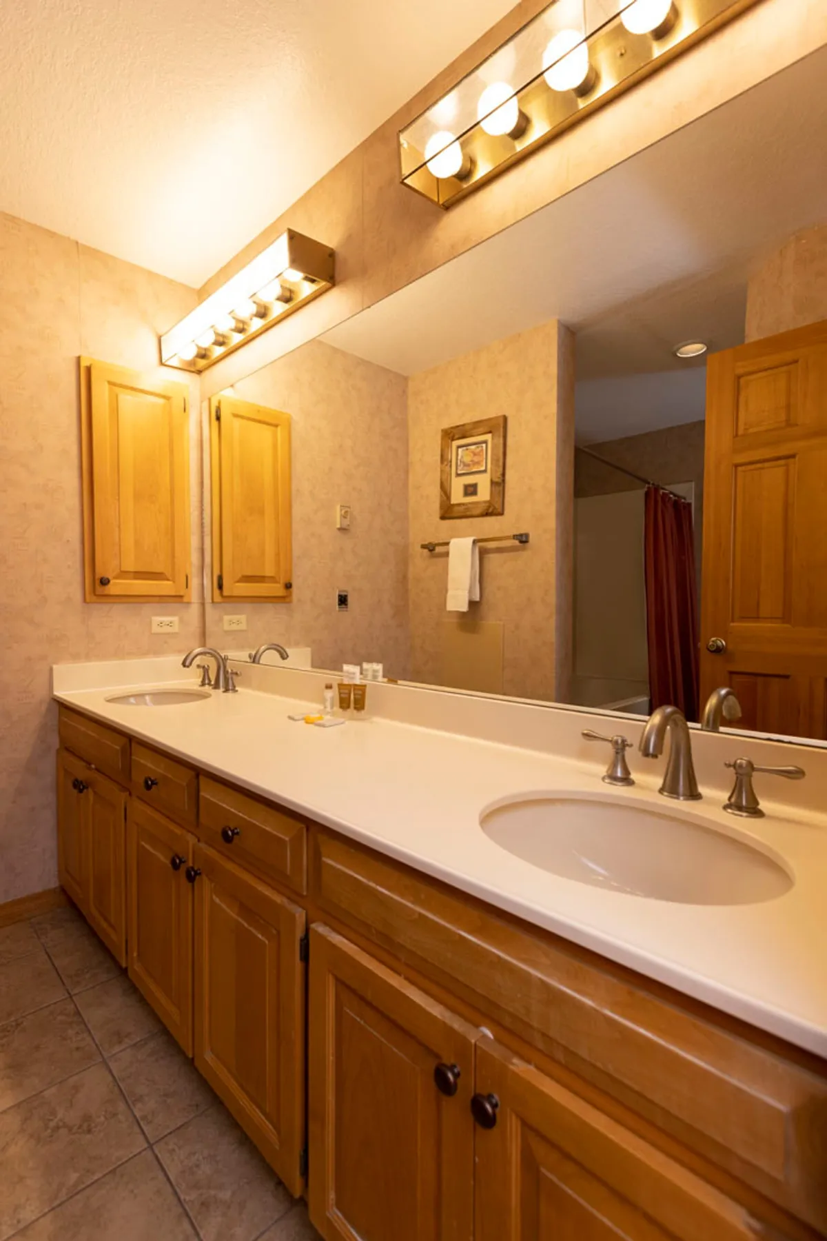 A spacious bathroom with double sinks, wooden cabinetry, modern light fixtures, and framed artwork. The organized, calm setting is ideal for morning routines and self-care, creating a seamless transition from activity to rest.