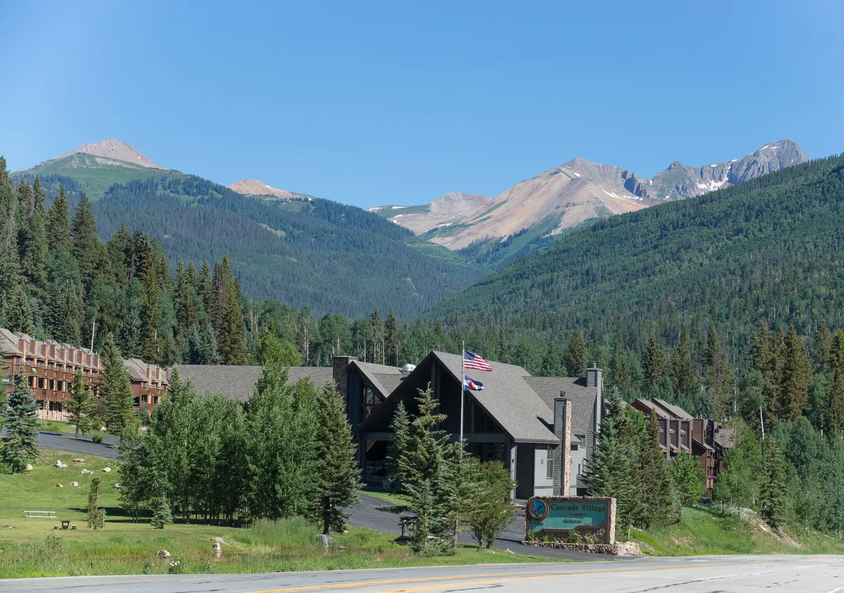The main building of Cascade Village stands against a dramatic mountain backdrop, surrounded by evergreens and clear signage. The scene exudes a sense of arrival and connection to nature, with visual cues to outdoor recreation and a welcoming resort atmosphere.