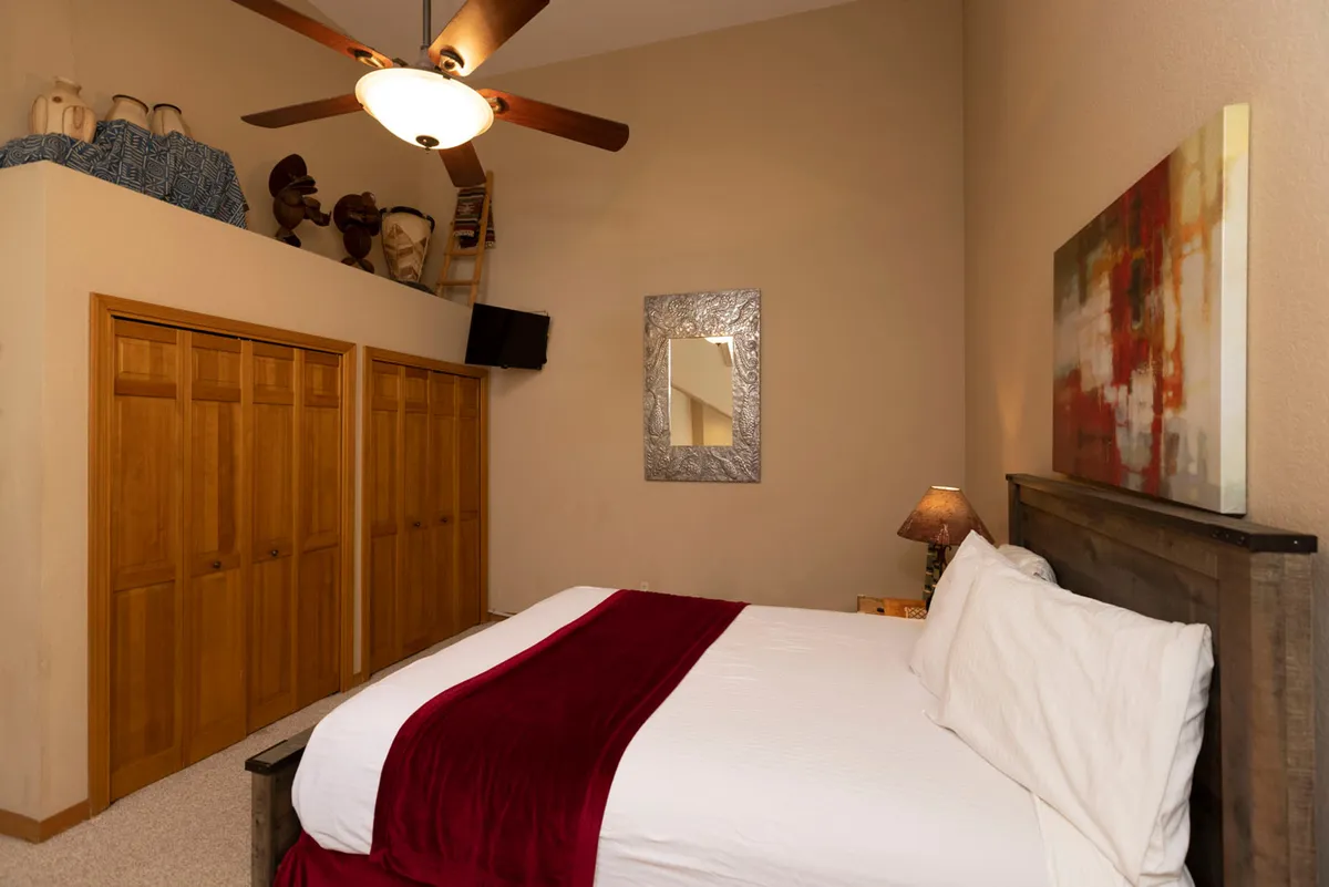A warmly decorated bedroom with a bed adorned with a red throw, wooden closet doors, decorative masks, and abstract artwork. The space exudes warmth and comfort, ideal for relaxing or reading after a day of adventure.