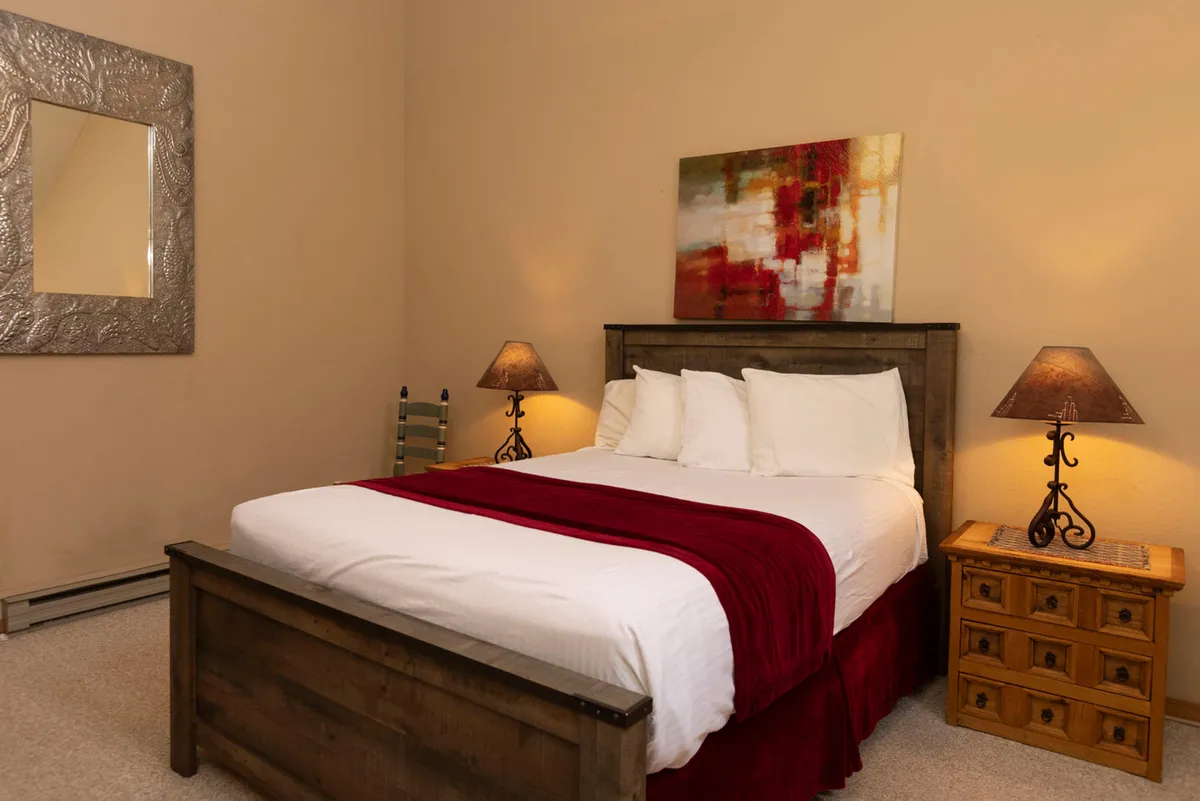 A cozy bedroom scene with a plush bed adorned with a red throw, flanked by soft lighting from bedside lamps and accented by abstract artwork above. The inviting setting suggests comfort and relaxation, perfect for quiet moments or winding down after a day exploring the mountains.