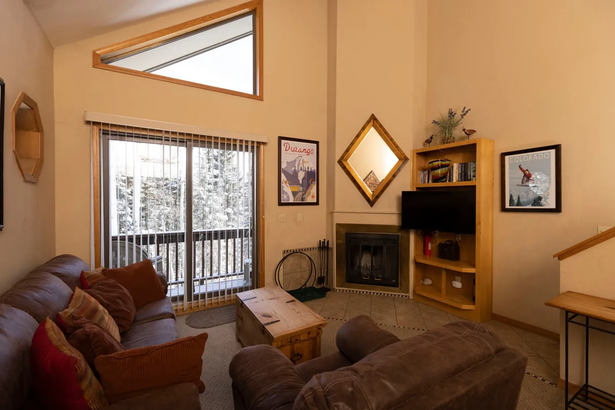 The image showcases a cozy living room with a stone fireplace as the centerpiece, flanked by large windows that let in natural light and frame the picturesque Colorado landscape. A plush sofa invites relaxation, while a wooden bookshelf and decorative mirror add warmth and charm to the space. The setup suggests a perfect spot for unwinding after mountain adventures, reading by the fire, or enjoying the serene views outside.