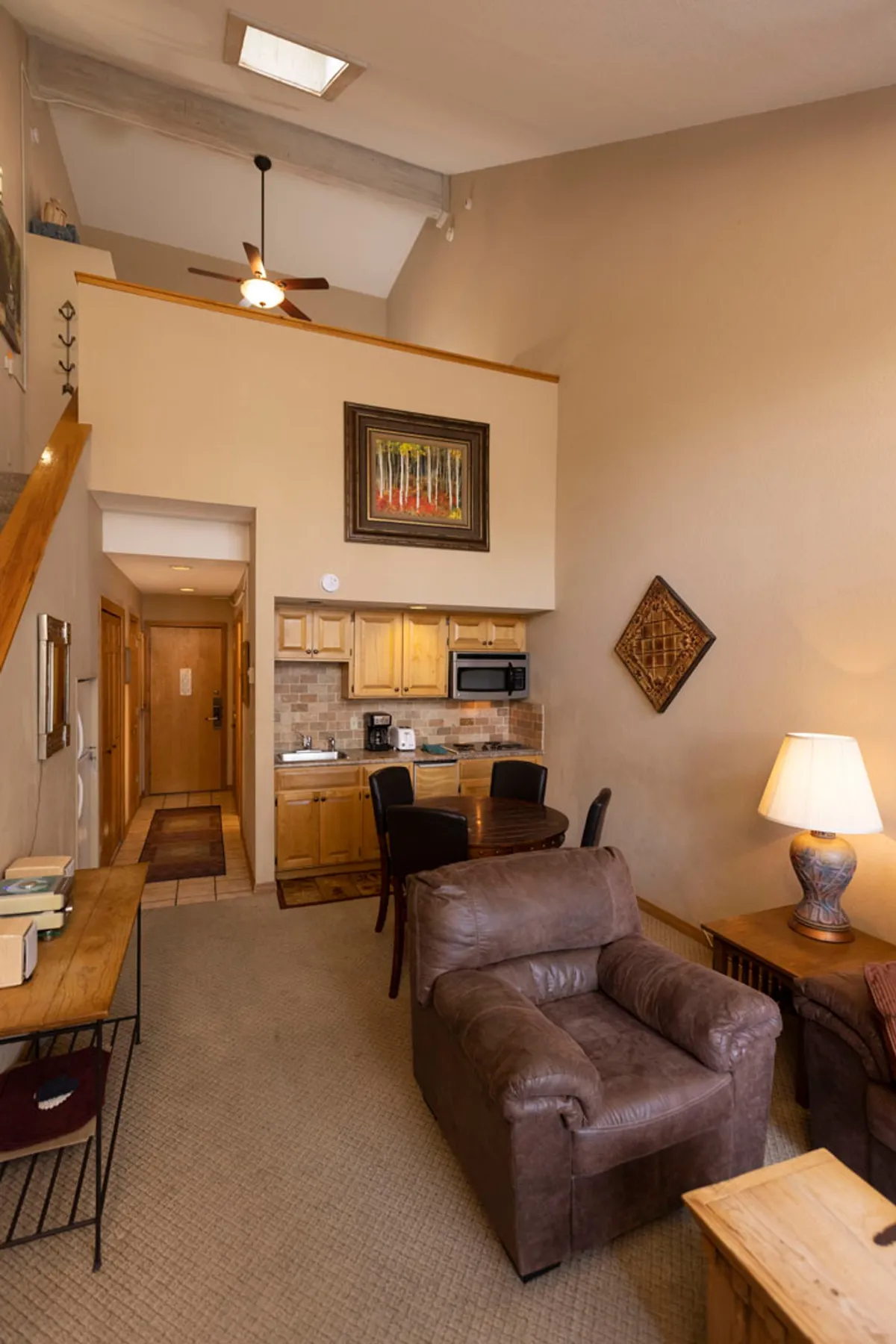 A welcoming interior with a leather armchair, wooden dining table, kitchen area, decorative wall art, and a lamp. The space is warm and inviting, ideal for relaxing and socializing.