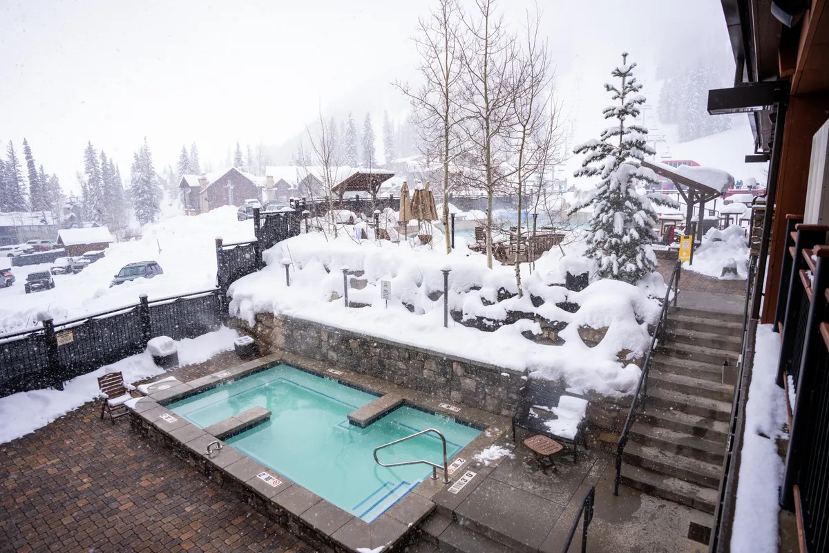 An outdoor hot tub nestled in a snowy, tree-lined landscape, with a stairway leading up, suggesting easy access. Steam rises gently, evoking warmth and relaxation amid the winter scenery.