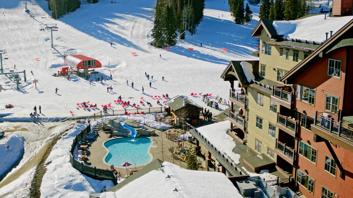 A lively winter scene showing expansive ski slopes, a ski lift transporting guests up the mountain, an outdoor pool steaming in the cold, the welcoming resort lodge, and people actively skiing. The setting is bustling with winter activity and framed by snow-covered terrain, highlighting the vibrant atmosphere of the resort.