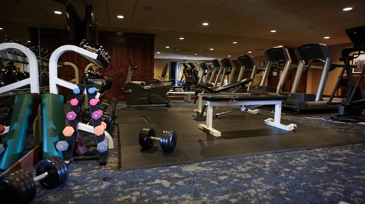 This image features an interior fitness room with dumbbells, treadmills, and an exercise bench. The space is well-lit and energetic, designed for group fitness or personal workouts.